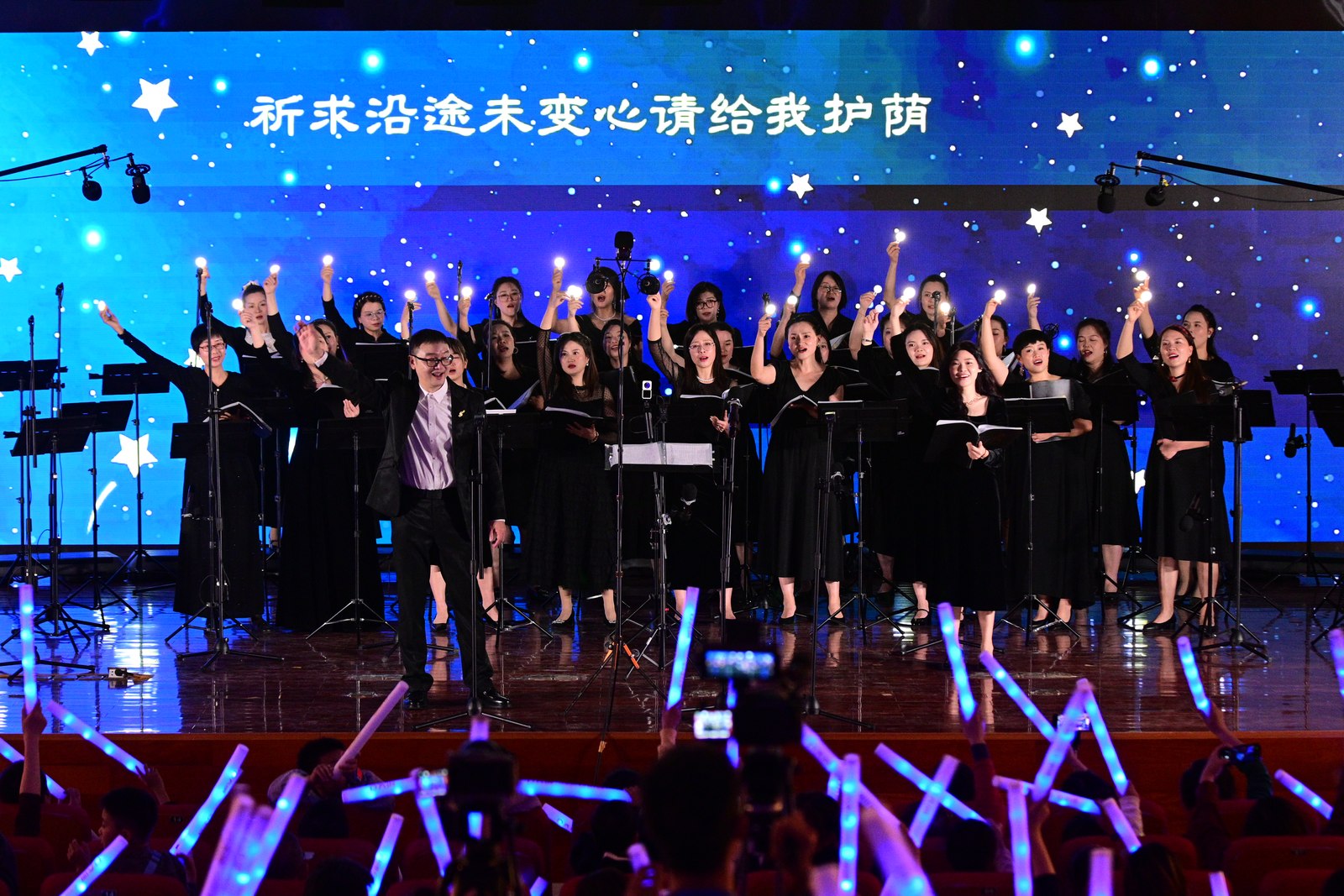Choir members in black concert dress raising their arms under blue stage lights