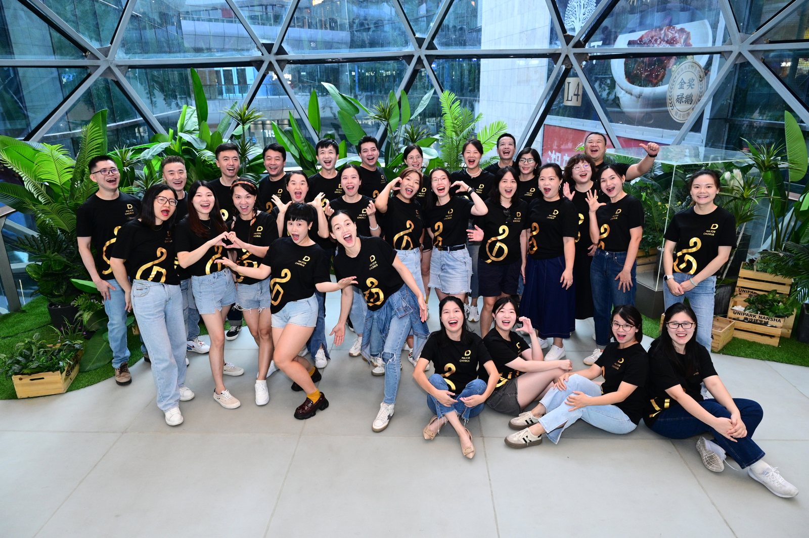 D Major Choir members in black choir shirts posing together by greenery and a glass wall